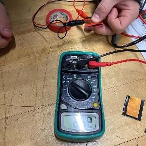 Closeup of a student’s hand squeezing a DIY FSR from copper sheet scraps and conductive foam along with the multimeter being used to measure the resistance value. There is a glare on the display that obscures the reading.