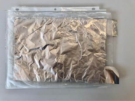 A photograph shows the stack of tin foil and wax paper together placed inside of a clear sheet protector so that the tin foil tails are sticking out of the sheet protector. 