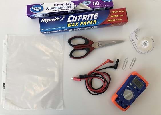 A photograph shows a clear sheet protector, a box of tin foil, a box of wax paper, scissors, clear tape, paper clips, black and red alligator clips, and a capacitance meter laying on a table. 