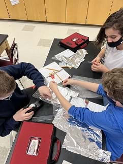 A photograph shows students around a table building a sensor. The student on the left is taping a piece of tin foil to the wax paper. One student on the right is viewing the other student on the right taping the other side of the tin foil to the wax paper.