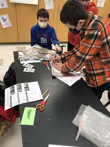 A photograph shows students around a table building a sensor. A student at the top of the photo sits in a chair and is grasping a piece of tin foil. A student is standing next to him watching. Another student to the right is using a marker to draw a line to cut out a tin foil piece for their sensor. 