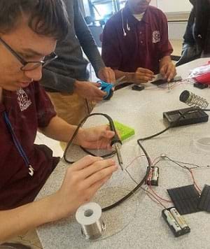 A photograph shows three students at a table; one is using a soldering iron and focused on building his solar charger.