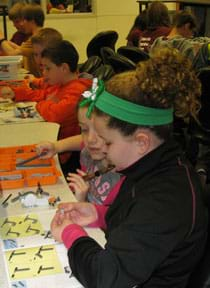 A photograph shows two girls working together at a lab bench, following picture instructions to put together pieces to make a LEGO robot. Nearby, other teams do the same.