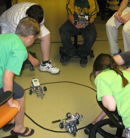 A photograph shows six students in a circle watching two LEGO EV3 robots travel an oval line on the floor made with black tape.