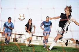 A photograph shows a girl kicking a soccer ball into a net goal as she is pursued by three other players.