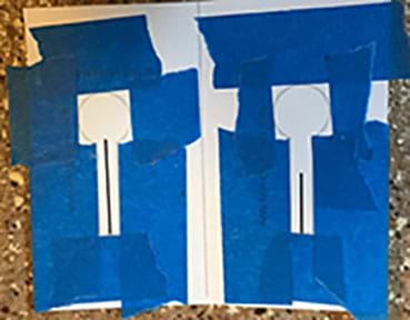 Photograph of a white card that has been folded down the middle and opened up to the inside. Blue painters’ tape has been applied to make stencils both sides. The shape is a vertical stem ending in a square.  
