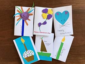 Photograph of six closed cards made by the author. Each one has one LED poking through. One card says “Happy Birthday” and has colored paper pieces of a candle glued to it. The LED is poking through where the flame of the candle is attached to the candle. Other cards are for Valentine’s Day (flower made of hearts and a mouse head made of hearts), Birthday (a cupcake with a lit candle), Congratulations (a fireworks, swirly pattern on a stick) and Bon Voyage (a sailboat).