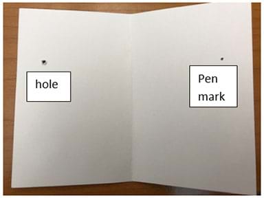 Photograph of an open piece of blank cardstock showing the inside where a hole has been poked through the left side with a pen point and a pen mark has been marked through the hole on the right side. 