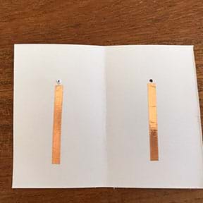 Photograph of the open piece of cardstock showing the inside where two pieces of copper tape have been placed vertically, one below the hole on the left inside cover and one below the pen mark on the right side.