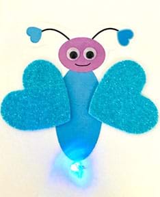 Photograph of card front shows Valentine’s Day bug made with blue glitter heart sticker wings and printed blue body, purple head with smile, antennae with blue hearts at their ends and “googly eyes” pasted on. A lit blue LED is at the tail.