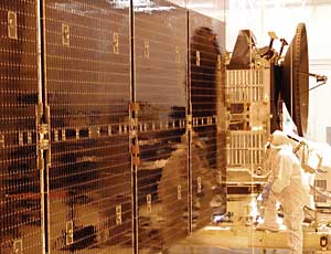 A photograph in a lab shows two people in white coveralls and hoods conducting pre-flight testing on the shiny, coppery solar array panels on the 27-foot long wings of the Dawn exploration spacecraft.