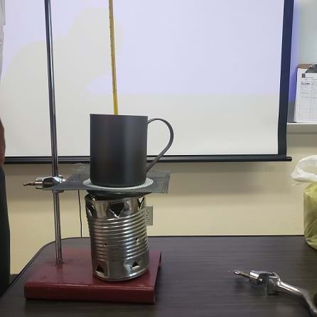 A tin can cookstove is sitting on a ring stand. A ring clamp is attached to the ring stand and wire mesh is sitting on the ring clamp. The ring clamp and wire mesh are sitting directly on top of the cookstove.  A steel mug is sitting on top of the wire mesh. 