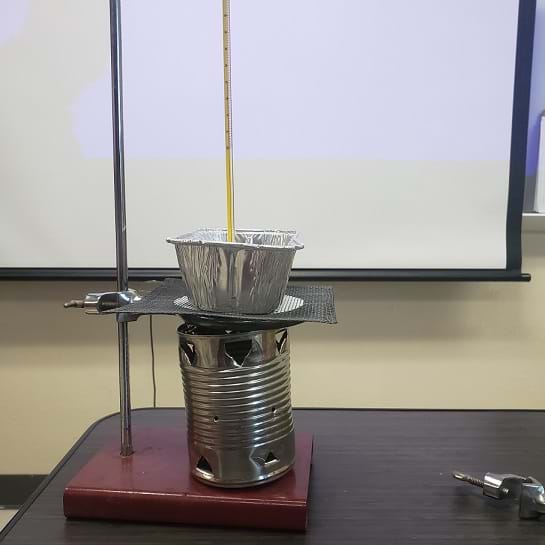 A tin can cookstove is sitting on a ring stand.  A ring clamp is attached to the ring stand and wire mesh is sitting on the ring clamp. The ring clamp and wire mesh are sitting directly on top of the cookstove.  An aluminum loaf pan is sitting on top of the wire mesh. 