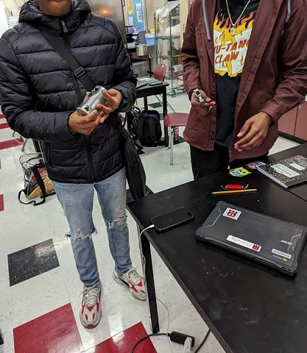 One student holding a tin can with holes in it standing next to a student holding a hammer. Both students are standing at a table/workstation.