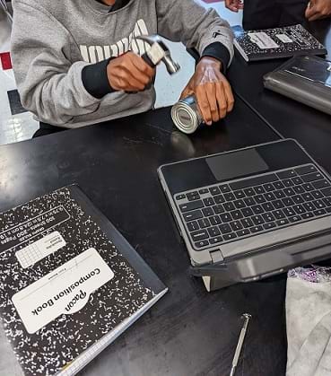 A student sitting at a table pounding a tin can with a hammer. A laptop and notebook are on the table.