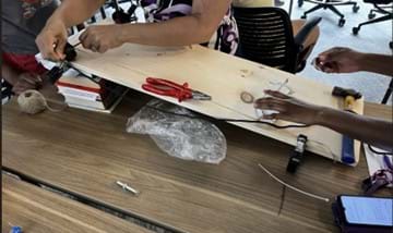 A photo showing students working to set up an inclined plane using a wooden plank propped on books, with multiple hands adjusting the black string and a white plastic cup (the load). Various tools and materials, including pliers, a hammer, and twine, are scattered nearby as they prepare or test the setup. A photo showing students working to set up an inclined plane using a wooden plank propped on books, with multiple hands adjusting the black string and a white plastic cup (the load). Various tools and materials, including pliers, a hammer, and twine, are scattered nearby as they prepare or test the setup.