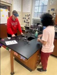 A photo of two students facing each other who appear to be chatting while working in a science lab. One is standing in front of a Bunsen Burner and ring stand setup with a beaker with a yellow solid inside, while the other is using a mortar and pestle. 
