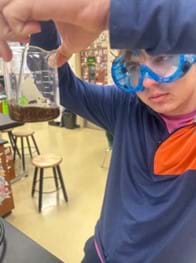 A photo of a student wearing blue goggles and holding up a beaker containing a thick brown fluid to inspect while he stirs.