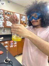 A photo of a student wearing blue goggles who is holding up a beaker containing a thick brown fluid and inspecting it while he stirs.