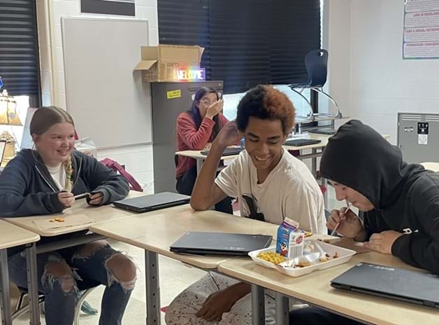 A photo showing three students sitting at a table in a classroom, two boys and one girl. One of the boys is attempting to suck up a Goldfish cracker with a straw while the other two laugh.