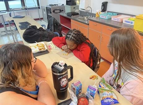A photo showing three girls sitting at a table in a classroom, two with their back to the camera and one looking down at the desk. One of the girls is attempting to move Goldfish crackers with her straw while the other two are watching.