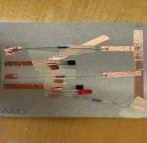 A photo of an AND gate built on a piece of cardboard from a cereal box cut to the size of a notecard. The gate is built using copper tape, three resistors, two transistors and an LED. There is also masking tape used to separate layers of the copper tape to prevent short circuits. There are two places in the circuit where gaps are left between pieces of copper tape to create the switches in their open states.