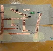 A photo of an OR gate built on a piece of cardboard from a cereal box cut to the size of a notecard. The gate is built using copper tape, three resistors, two transistors and an LED. There is also masking tape used to separate layers of the copper tape to prevent short circuits. There are two places in the circuit where gaps are left between pieces of copper tape to create the switches in their open states.
