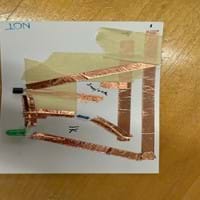 A photo of a NOT gate built on a piece of cardboard from a cereal box cut to the size of a notecard. The gate is built using copper tape, three resistors, two transistors and an LED. There is also masking tape used to separate layers of the copper tape to prevent short circuits. There are two places in the circuit where gaps are left between pieces of copper tape to create the switches in their open states.