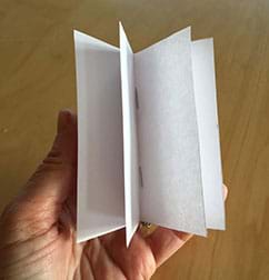 A hand holds a small booklet made of blank white paper with its four pages spread apart so you can see each one. The booklet is composed of two, folded 4 x 4-inch sheets. You can see two vertical staples along the inner center fold of the booklet.