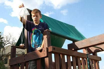 Image with a boy standing at the top of a slide in a wooden playground.  He is holding his parachute attached to a toy over the end of the wooden railing.