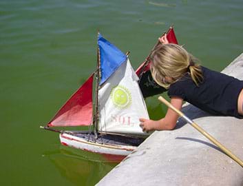 A small girl leaning over a concrete ledge to place her small homemade sailboat into water.