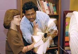 Image of a male Air Force engineer standing with a 5th grade girl holding a hand-made wind turbine with electrical wires attached to it.  They are holding the wind turbine in front of a fan to test its energy output.