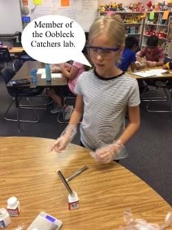 A female fifth grader stands at a table. She is wearing protective gloves and safety glasses. The table contains scoopulas and NaNupp Dust in a white plastic bottle. Students are working behind her at their lab tables. A dialogue bubble states “Member of the Oobleck Catchers Lab.”