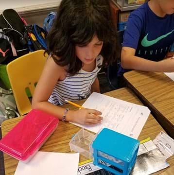 A photograph of a girl evaluating a 3-drawer organizer. The girl is recording her observations on a piece of paper using a pencil.