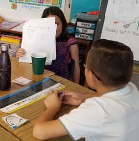 A photograph of a girl raising her paper to show a boy her ideas. The boy is holding “talking chips” in his hand.