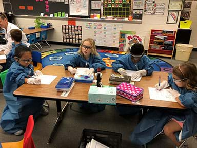 Four children around a lab table. They are sharing a microscope and documenting what they see through the lens.