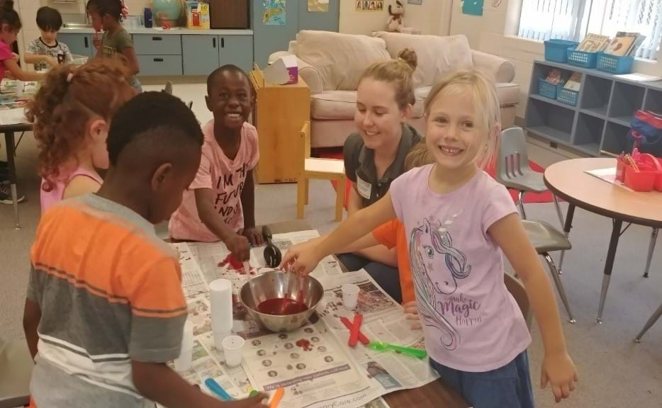 Four students and one adult volunteer around a small table stirring a red liquid in a metal bowl.