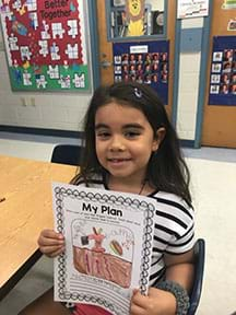 A female student displays her plan for the red wiggler’s habitat. Her picture includes dirt with worms below the surface, an apple core, and paper. She has labels on her picture. Below the picture is writing about her model habitat.