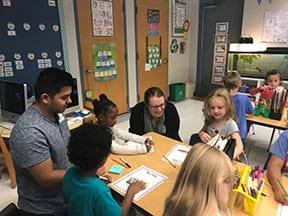 Two University of Florida graduate students in the Material Sciences and Engineering department work alongside a group of students at a table. The students are drawing and writing the details of their red wiggler’s model habitat. You can see two students in the background working on the same assignment.