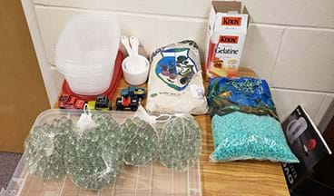 An image of plastic containers, marbles, toy trucks, gelatin packets, sand, pebbles and measuring cups sit on a table. 