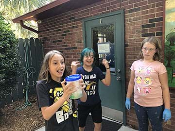 Three students stand outside a school and shake a container filled with detergent and mustard-stained cloth. 