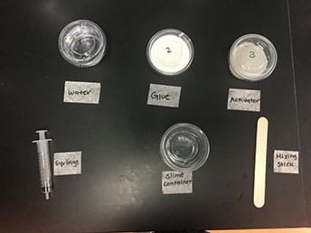 An image of a small cup of water, glue, activator along with a syringe, slime container, and stirring stick. 