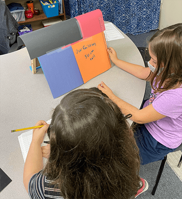 One student is measuring their construction paper building with a ruler, which reads thirty-six centimeters. Another student is recording their data in a notebook. 