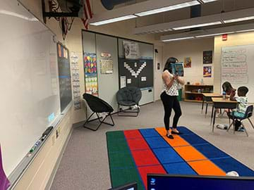 A teacher stands in front of a elementary school classroom reading a book.