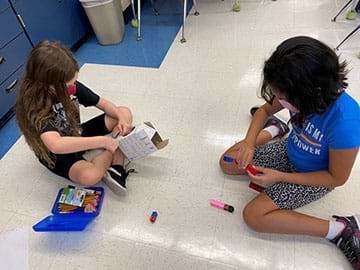 Two students work to construct a personal snack holder. One is using materials from a tissue box and the other is using connecting blocks.