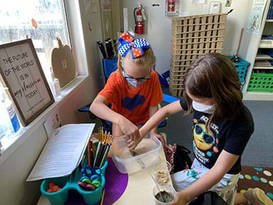 In a classroom, two students build a prototype seawall in a plastic container.