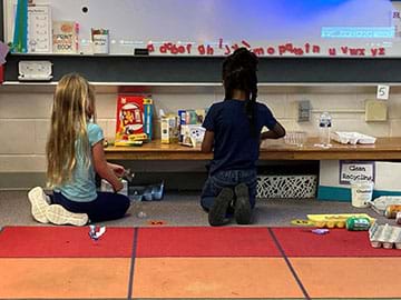 A girl and a boy work at a bench creating inventions using recycled plastic water bottles, egg crates, and paper food boxes.  
