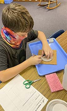 A student is tracing the top of an oatmeal container on a square of cork.