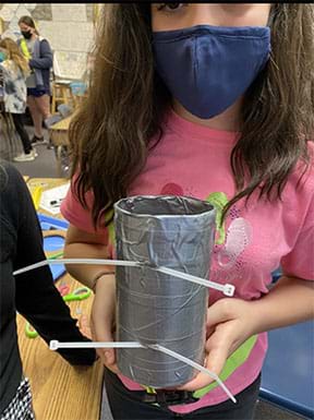 A student holds up a final design of her group’s water bottle holder before attaching it with two zip ties to the leg of a desk.