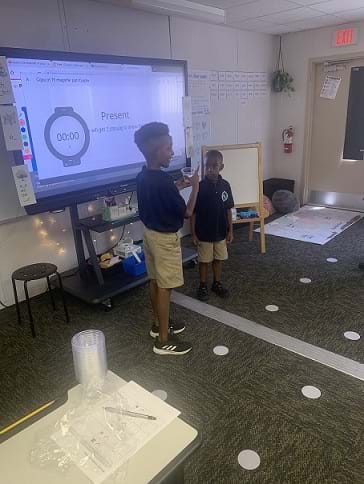 Two students stand in front of a smartboard and present their findings to their classmates.